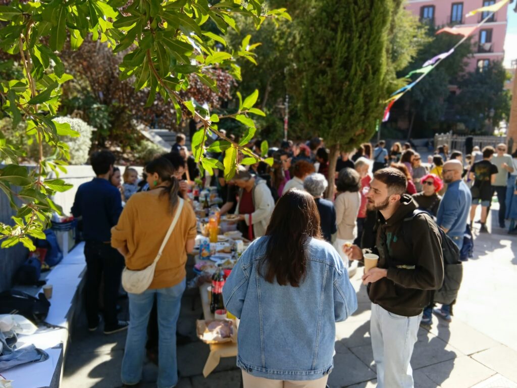 Vecinas y vecinos compartiendo comida en una mesa larga en la Plaza del Dos de Mayo